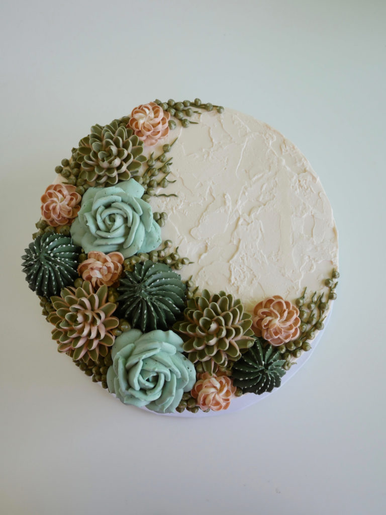 Overhead shot of buttercream covered round cake with ivory icing and a crescent shaped bouquet of buttercream succulents in muted green and coral