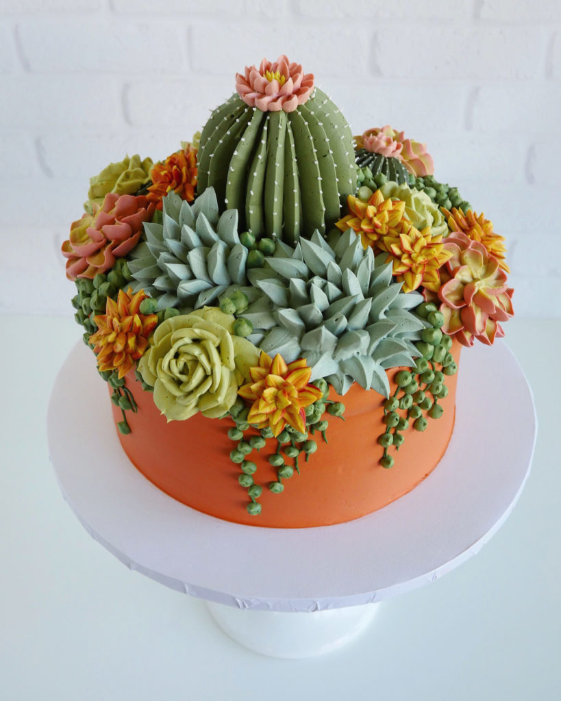 Overhead shot of tones of muted green and orange buttercream piped succulents and cactus on top of a round terra cotta colored cake